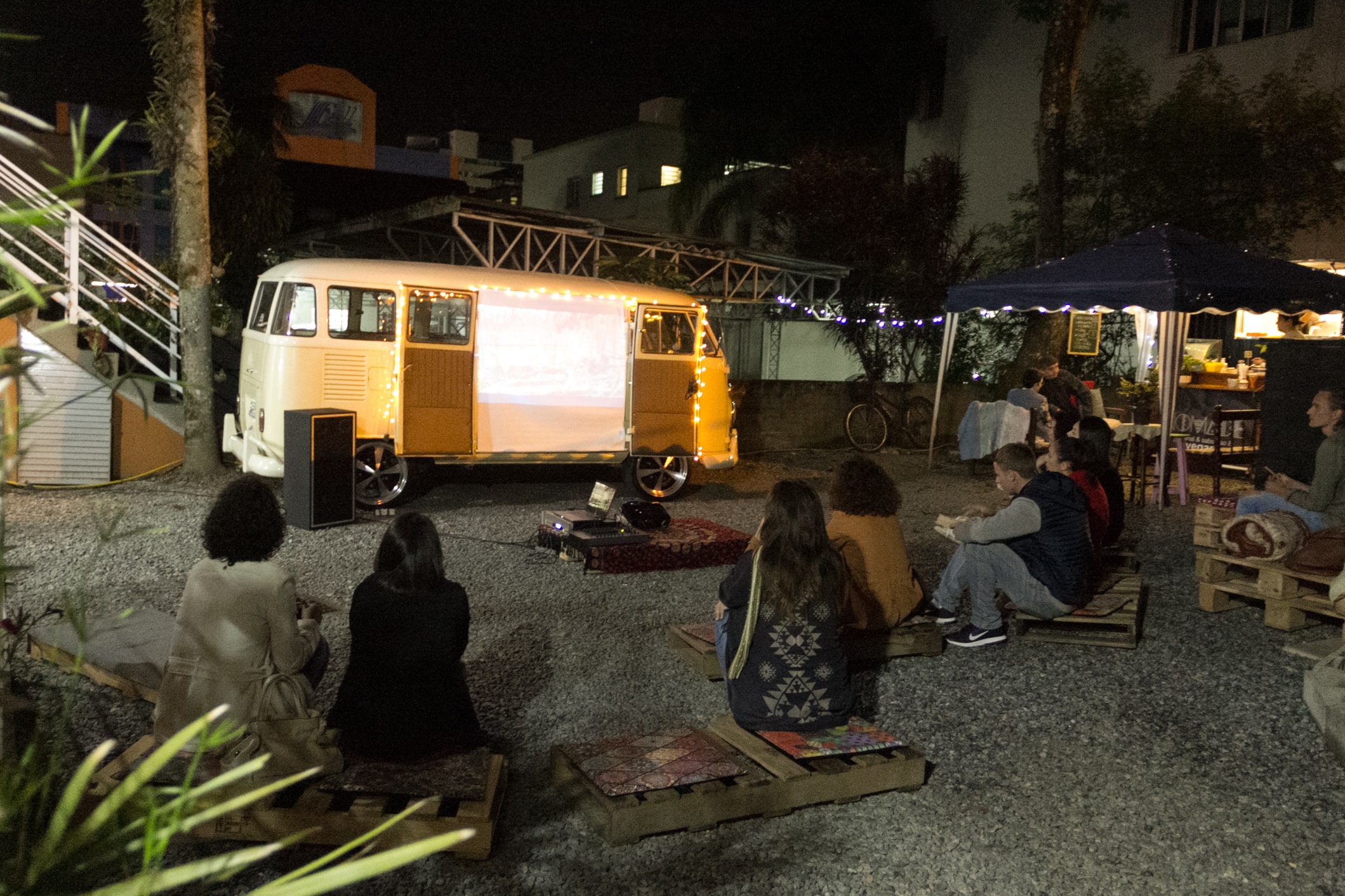 Em um local aberto pessoas sentadas em pallets de madeira no ch\xe3o. \xc9 noite e elas assistem a um filme projetado em uma tela fixada na porta de uma kombi amarela.