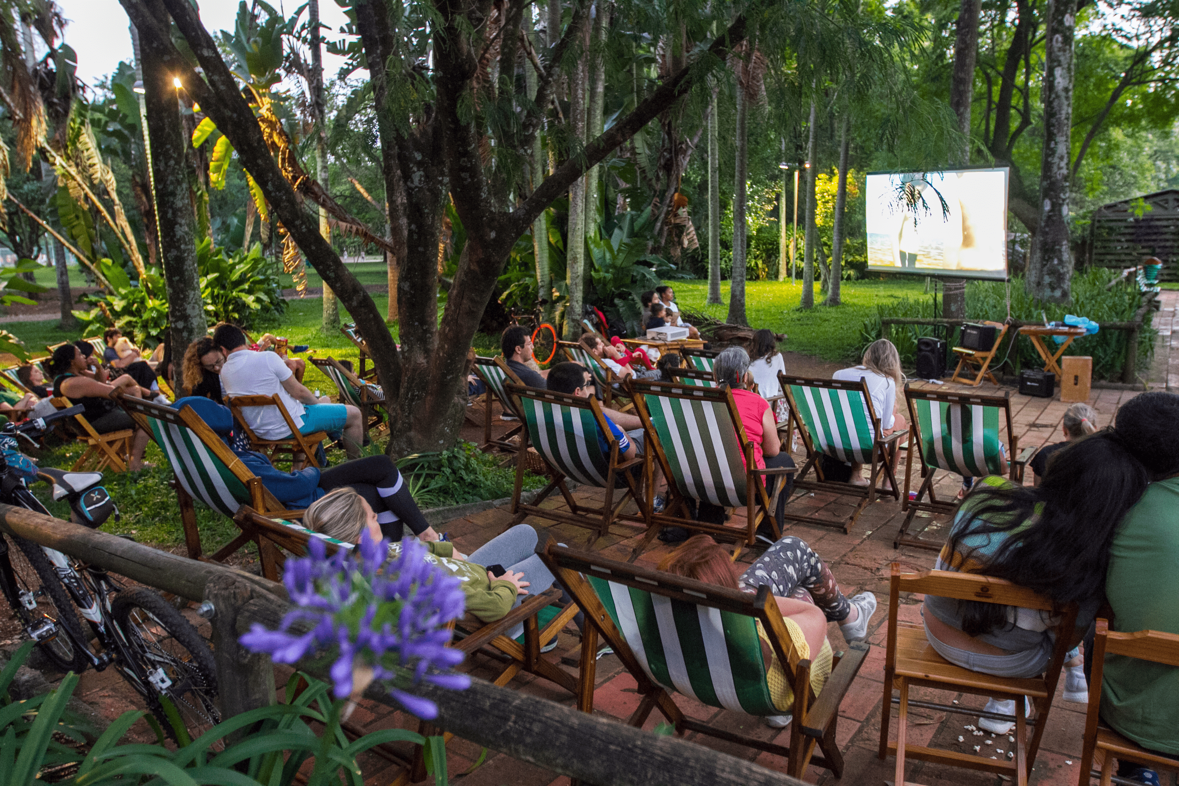 Em um jardim gramado e com \xe1rvores, no final de tarde, pessoas est\xe3o sentadas em cadeiras de praia e assistem a um filme projetado em uma tela fixada em uma das \xe1rvores do espa\xe7o.
