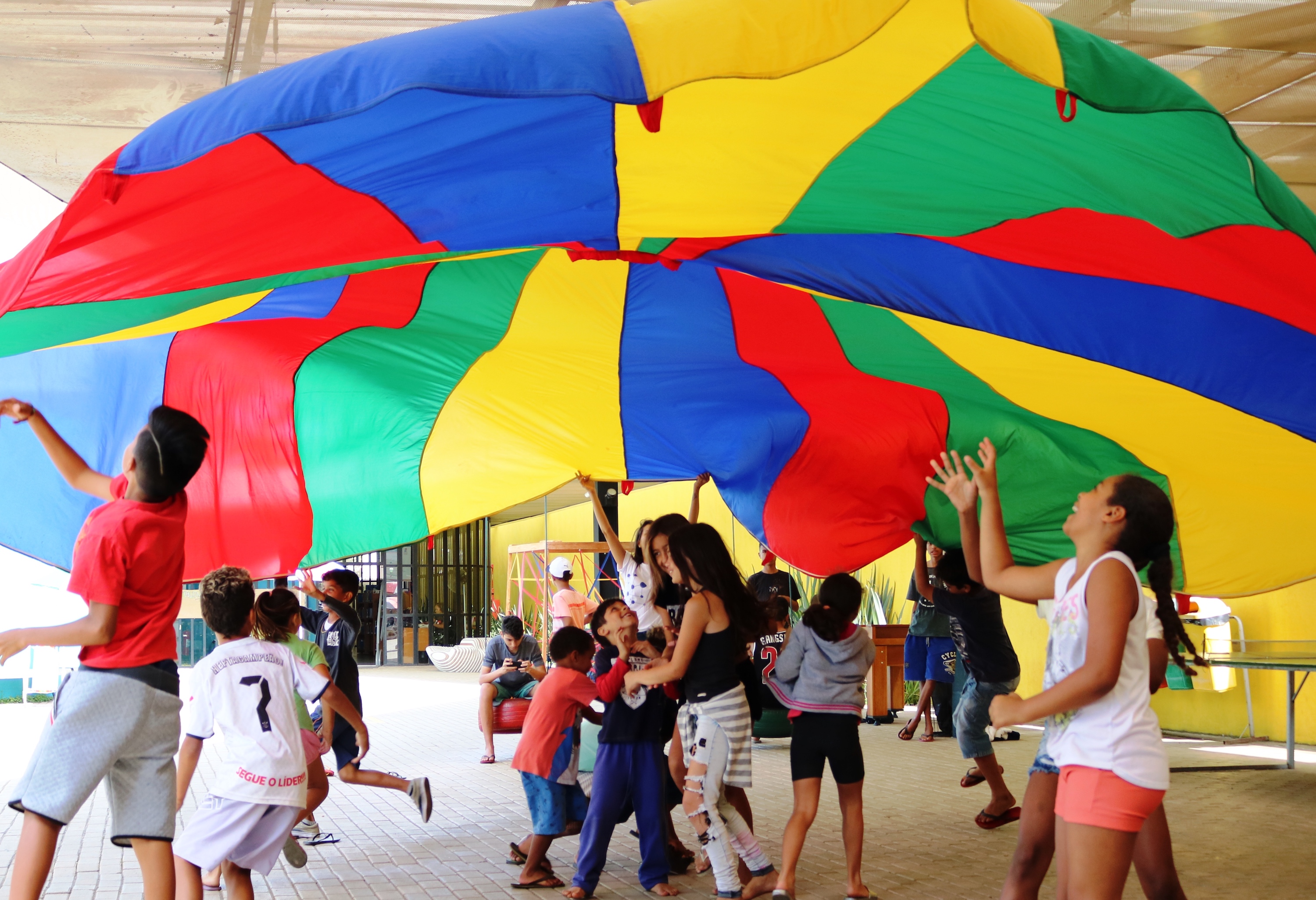 Crian\xe7as e adolescentes brincam embaixo de uma grande tenda colorida, amarela, vermelha, azul e verde.